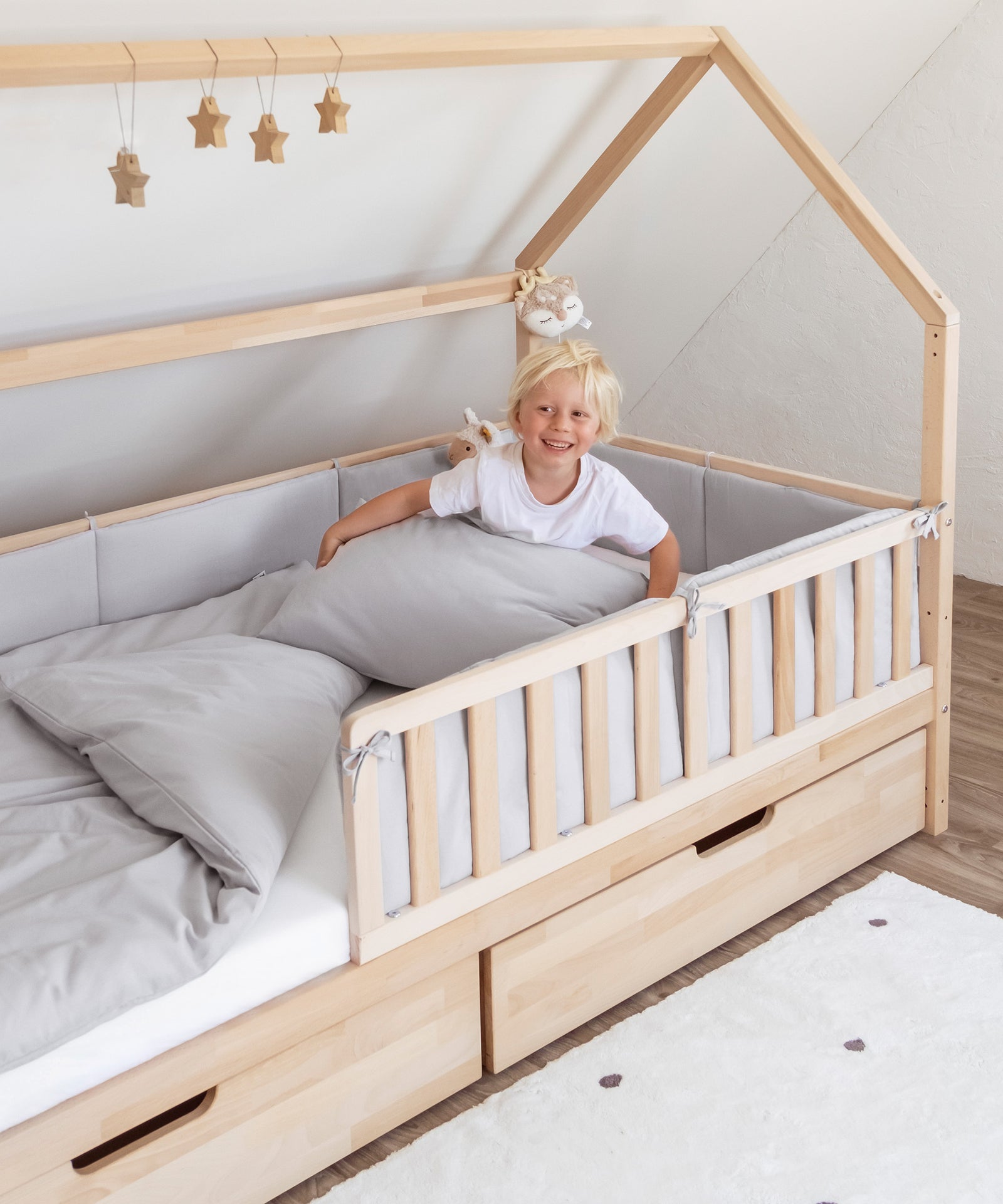 Waschbar & pflegeleicht*Die Bettumrandung ist bis 40 °C in der Waschmaschine waschbar und kann bei niedriger Temperatur im Trockner getrocknet werden.|Kind sitzt lachend in Hausbett mit Bettumrandung in Hellgrau.