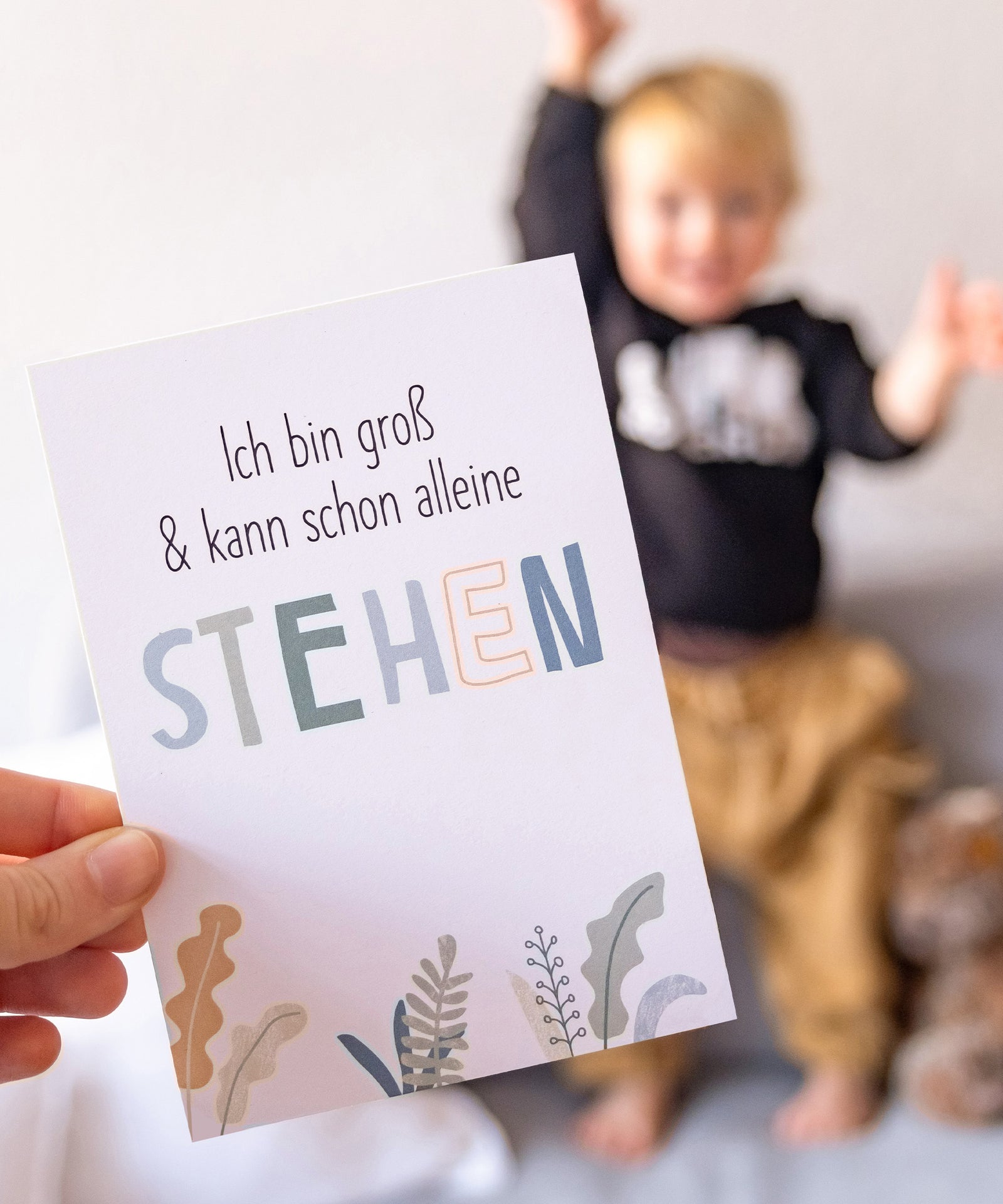 Besondere Momente festhalten*Mit den Meilensteinkarten kannst du wertvolle Erinnerungen an besondere Momente im ersten Lebensjahr deines Babys festhalten.|Karte mit Spruch „Ich bin groß und kann schon ganz alleine stehen“ mit stehendem Baby im Hintergrund.