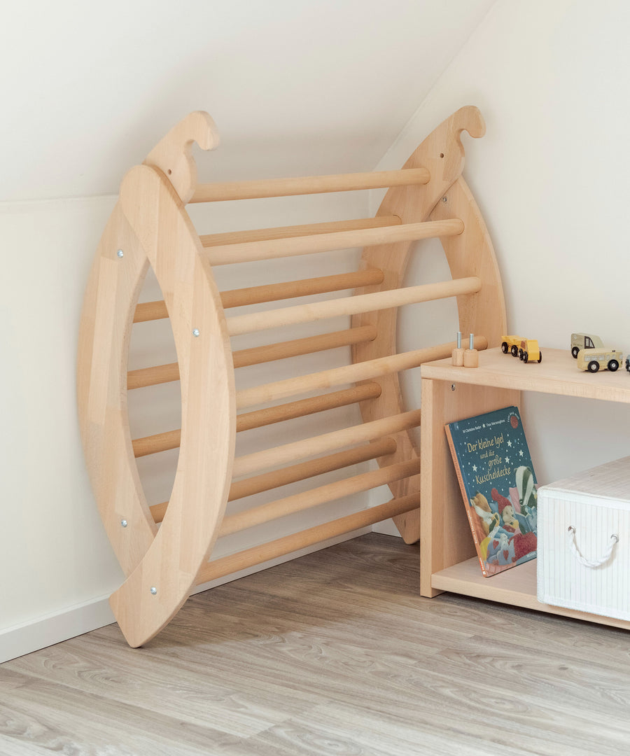 Zusammenklappbar*Der Kletterbogen kann mit wenigen Handgriffen platzsparend zusammengeklappt werden.|Zusammengeklappter Kletterbogen steht aufgeräumt neben Spielregal.