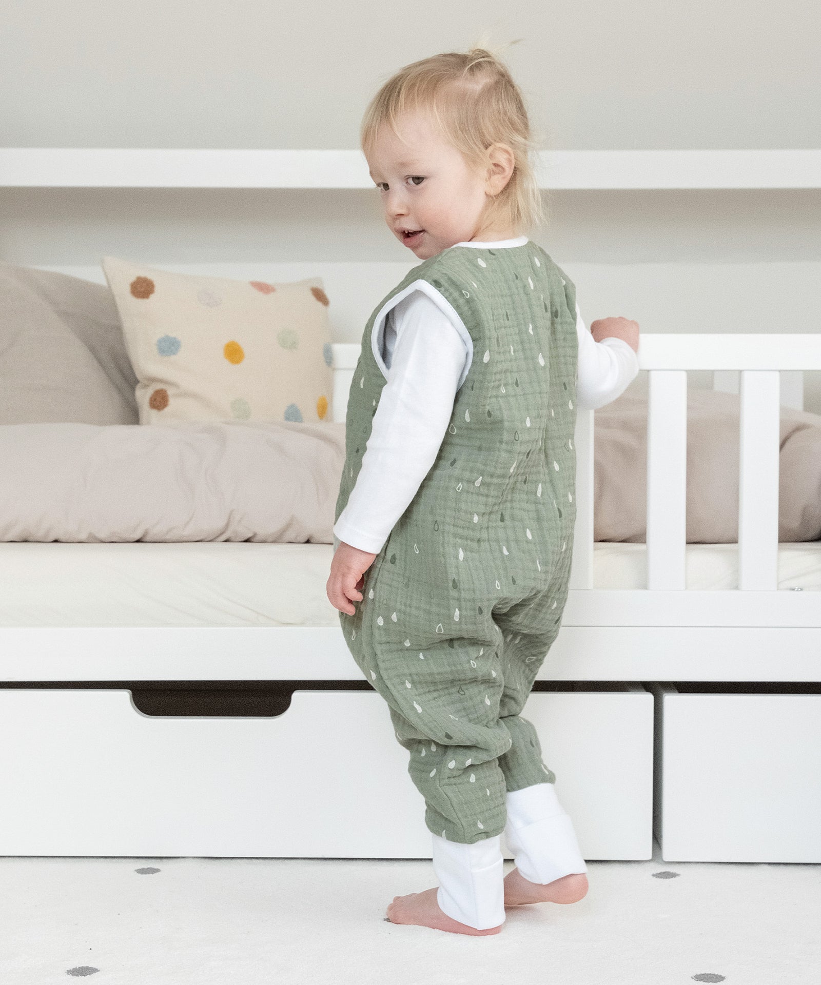 Mädchen mit Schlafsack aus Musselin steht an Kinderbett.