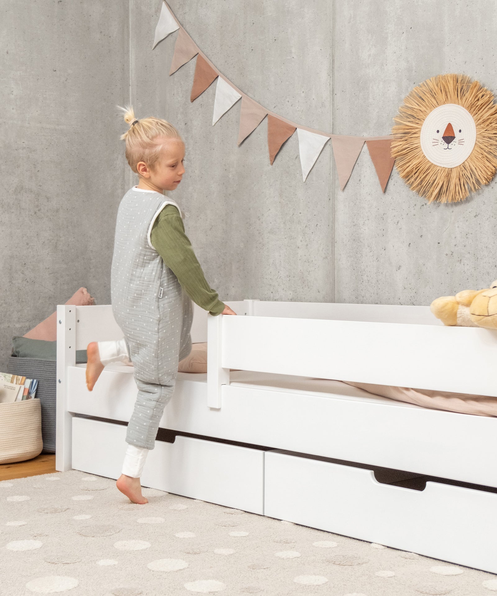 Abnehmbarer Rausfallschutz*Dank des Rausfallschutzes schlafen kleinere Kinder sicher. Er kann flexibel beidseitig montiert und leicht abgenommen werden.| Kind lehnt an Kinderbett Buche weiß.