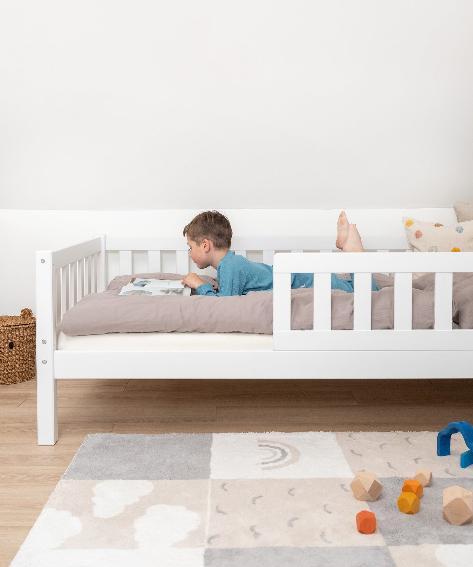 Flexibler Rausfallschutz*Der Rausfallschutz bietet deinem Kind Sicherheit. Er lässt sich beidseitig montieren und bei Bedarf auch ganz abnehmen.|Junge liegt in Kinderbett Kiefer Weiß mit Rausfallschutz und schaut ein Buch an.