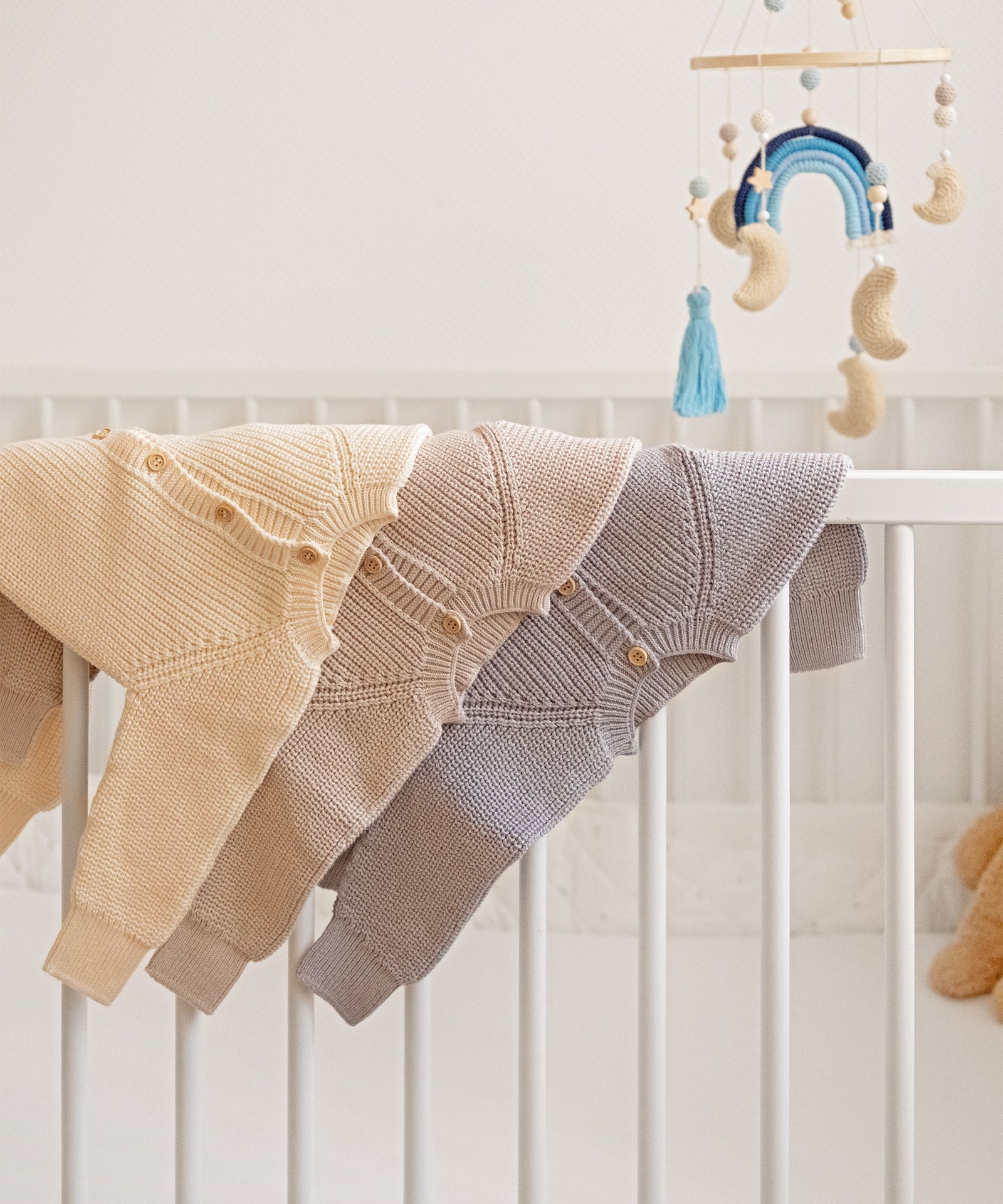 Strickoveralls in Beige, Natur und Hellgrau aus Bio-Baumwolle, dekorativ über der Babybett-Stange aufgehängt.